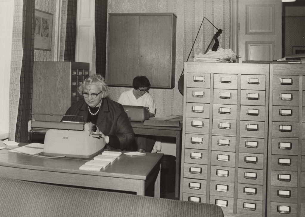 Tyra Petterson och Toini Koistinen skriver vistexter och registreringskort p&aring; skrivmaskin i stora arbetsrummet.