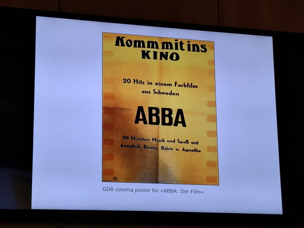 Affisch för filmen ABBA: Der Film på en presentationsskärm. Affischen är gul med svart text på tyska och lyfter fram ABBA samt information om filmen.