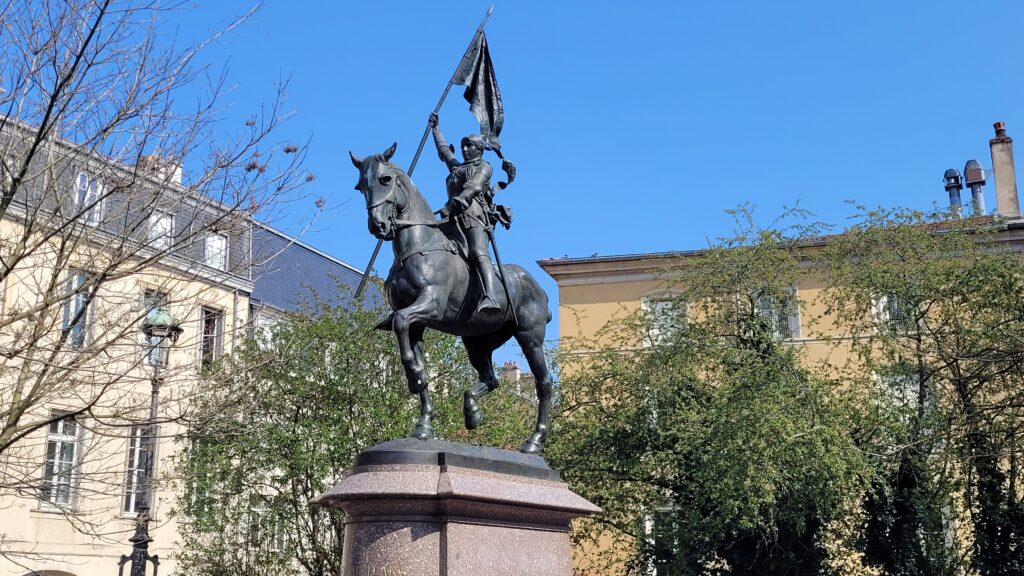 Ryttarstaty av Jeanne d’Arc i Nancy. Hon sitter till häst och håller en fana högt, med byggnader och träd i bakgrunden under klarblå himmel.