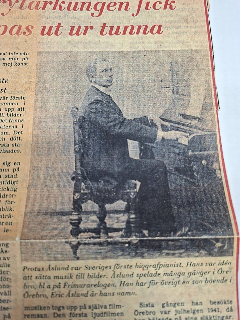 Svartvitt fotografi av biografpianisten Protus Åslund sittande vid ett piano. Han är klädd i mörk kostym och vänder sig mot kameran med händerna på tangenterna. Bilden är återgiven i en tidningsartikel.