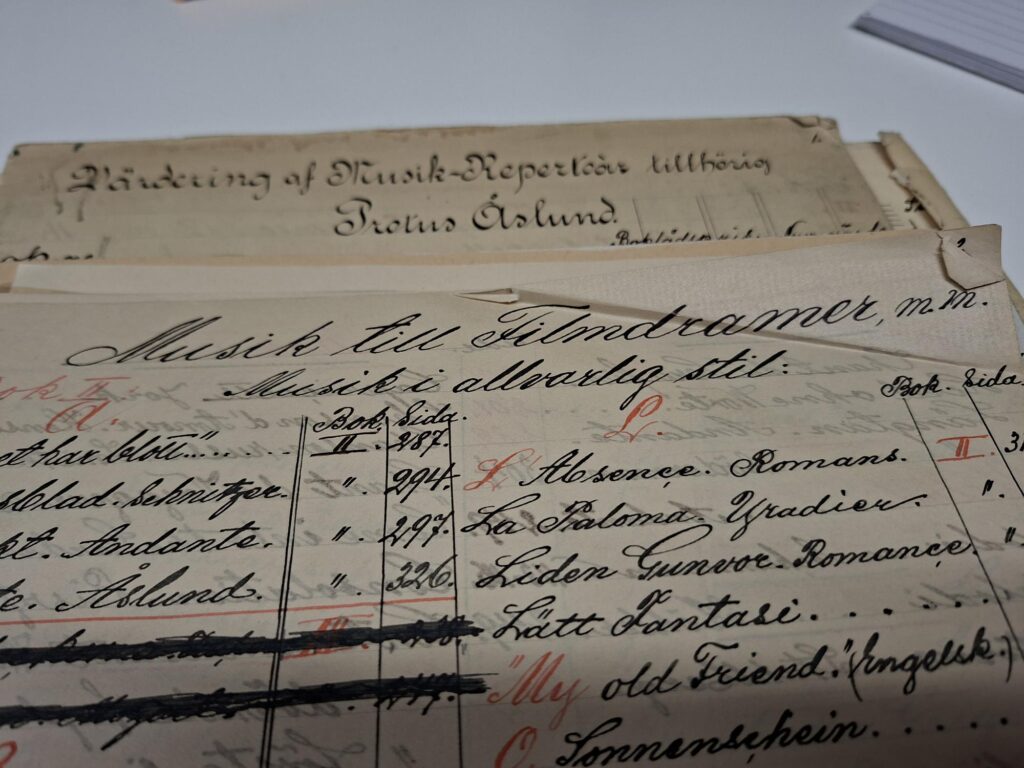 Närbild av handskrivna registerblad ur biografpianisten Protus Åslunds musikrepertoar. På bladen finns titlar till stumfilmsmusik och hänvisningar till nothäften, skrivna med svart bläck och kompletterade med anteckningar i rött. I bakgrunden syns ett omslag med texten "Förteckning af Musik-Repertoar tillhörig Protus Åslund".